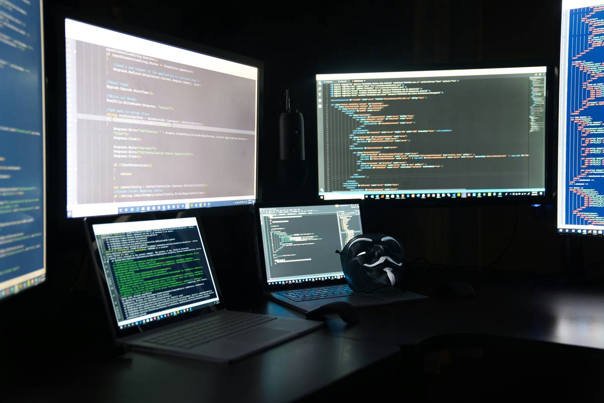 A computer work station featuring 2 laptops and multiple monitors of computer code with an anonymous (Anon) Guy Fawkes mask laying on one of the laptops.