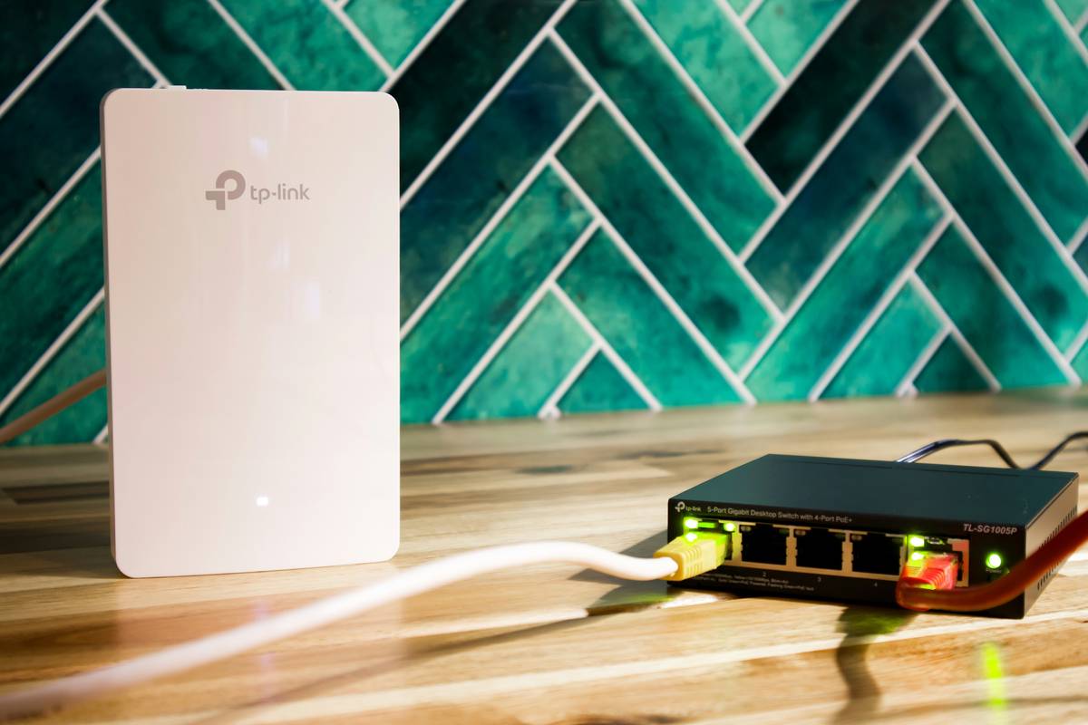 A contemporary networking setup is displayed on a polished wooden surface, featuring a white TP-Link WiFi access point paired with a black 5-port PoE gigabit switch. The scene is accentuated by a vivid, geometric green tile backdrop, creating a fresh and professional atmosphere. The glowing Ethernet ports and organized cabling emphasize reliable connectivity and advanced technology, making this image ideal for illustrating concepts like smart home networking, office IT infrastructure, or modern internet solutions.