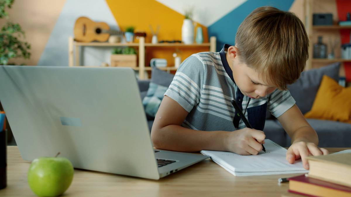happy child using laptop for education writing in notebook doing homework sitting at desk in apartment. Technology and lifestyle concept.